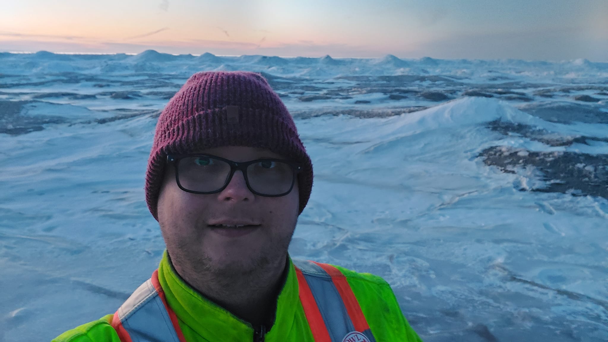 A man wears a red toque and yellow high visibility winter coat, standing in front of a landscape that looks like frozen waves, more of the landscape is visible, a little streak of water is visible in the distance